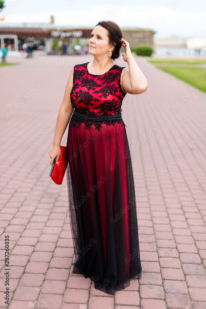 Brunette caucasian model in floor-length black and red dress corrects hair on street.
