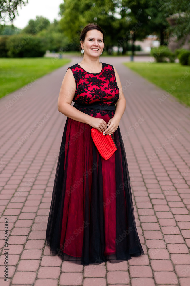 Brunette caucasian model in floor-length black and red dress with embroidery in park.
