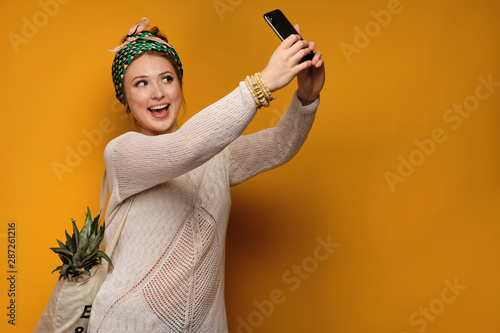 Fotografi Girl in a light sweater and knotted a colored head scarf with a pineapple in a bag and smiling happily makes selfies