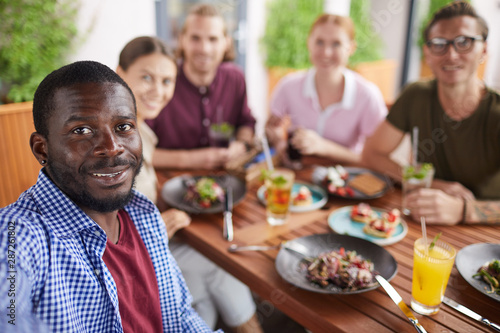 Multi-ethnic group of people posing for selfie photo while enjoying party dinner in cafe together  focus on smiling African-American man in holding camera in foreground  copy space