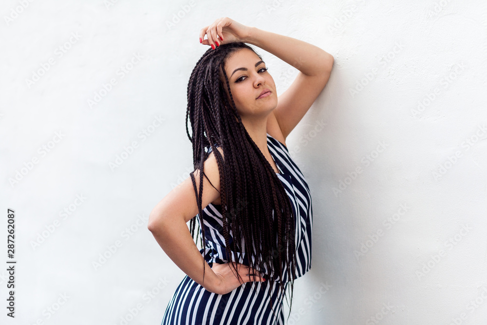 Portrait of serious young brunette woman with black afro american dreadlocks hairstyle in striped dress posing and looking at camera . indoor studio shot on white wall. Portrait of serious young brunette woman with black afro american dreadlocks hairstyle in striped dress posing and looking at camera . indoor studio shot on white wall.