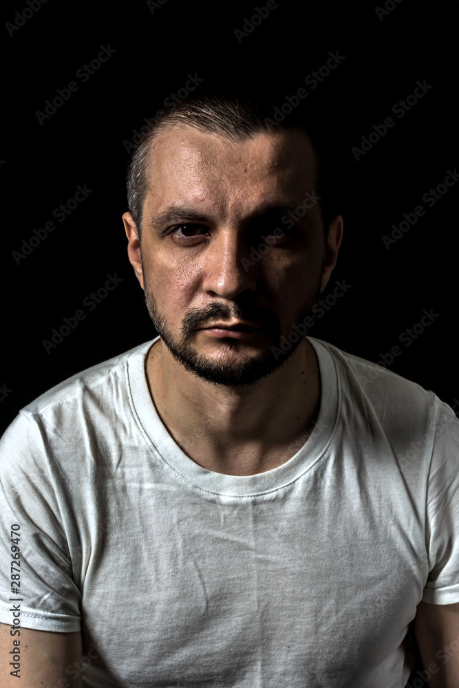 2019.05.19, Moscow, Russia. Portrait of a young serious man sitting on black background and looking to camera. Man's portrait in the dark.