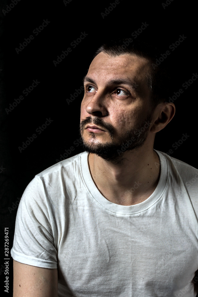 2019.05.19, Moscow, Russia. Portrait of a young serious man wearing white T-shirt sitting on black background and looking up away. Man's portrait in the dark.