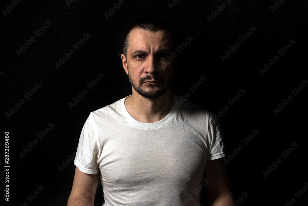 2019.05.19, Moscow, Russia. Portrait of a young serious man sitting on black background and looking to camera. Man's portrait in the dark. 2019.05.19, Moscow, Russia. Portrait of a young serious man sitting on black background and looking to camera. Man's portrait in the dark.