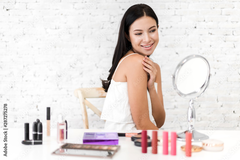 Smiling young beautiful woman fresh healthy skin looking on mirror with makeup cosmetics set at home.facial beauty and cosmetic concept Smiling young beautiful woman fresh healthy skin looking on mirror with makeup cosmetics set at home.facial beauty and cosmetic concept
