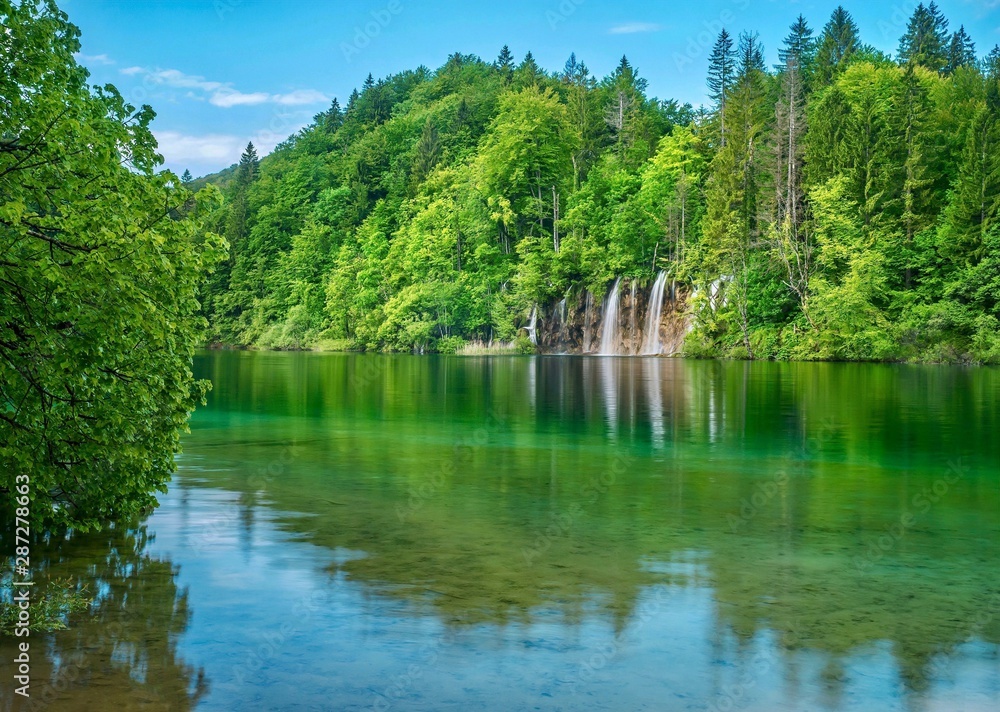 Fototapeta premium A beautiful summer nature scene in the forest, with waterfalls and trees reflected in a calm lake. Plitvice Lakes National Park, Croatia.