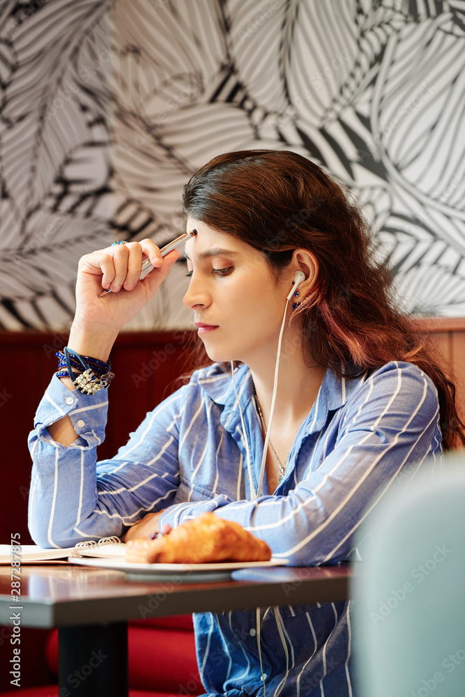 Young pensive female student sitting at cafe table, listening tin music and doing homework for tomorrow class Young pensive female student sitting at cafe table, listening tin music and doing homework for tomorrow class