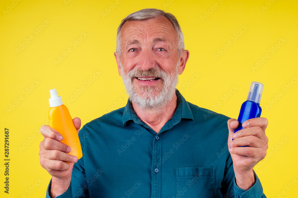 Senior man choosing sun protection cream between two spray bottle in studio yellow background Senior man choosing sun protection cream between two spray bottle in studio yellow background