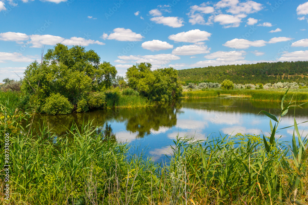 The beautiful calm river with green trees on the shore and white clouds on the sky