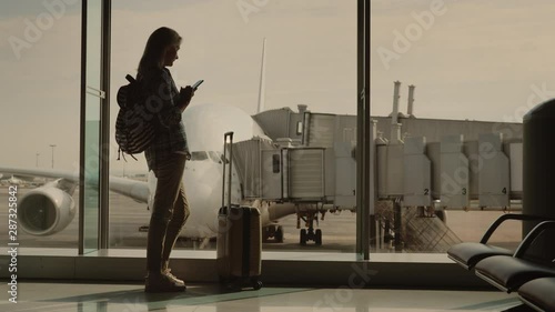 Wallpaper Mural A young woman with a backpack and a suitcase on wheels looks out the window at the plane and uses a smartphone Torontodigital.ca