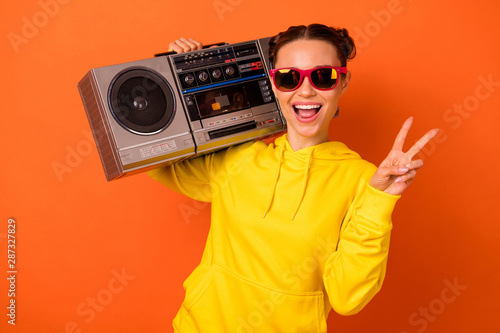 Portrait of excited girl having top-knot making v-signs holding boombox isolated over orange background
