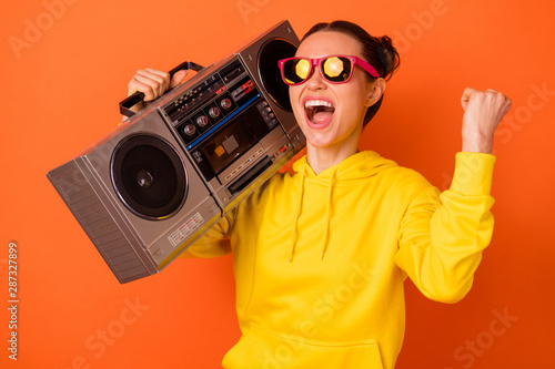 Portrait of cool teenager holding boombox shouting favorite song dressed eyewear eyeglasses raising fists isolated over orange background