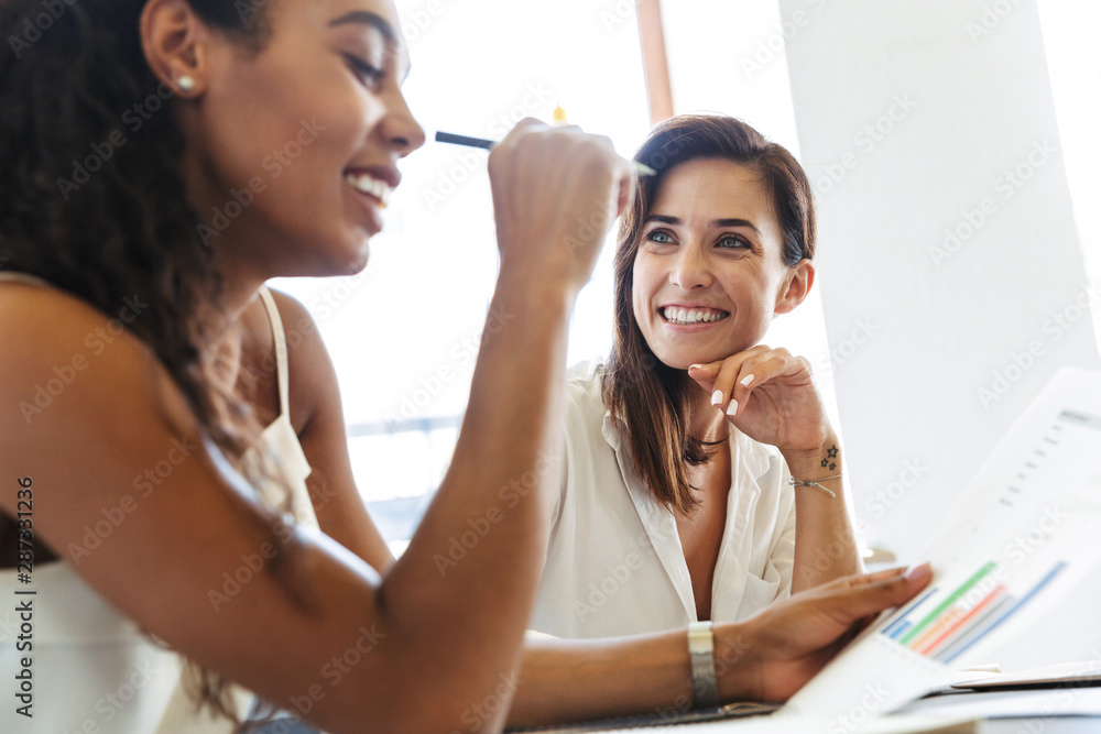 Cheerful happy positive young women colleagues at workplace indoors in office work with paper documents talking with each other. Cheerful happy positive young women colleagues at workplace indoors in office work with paper documents talking with each other.