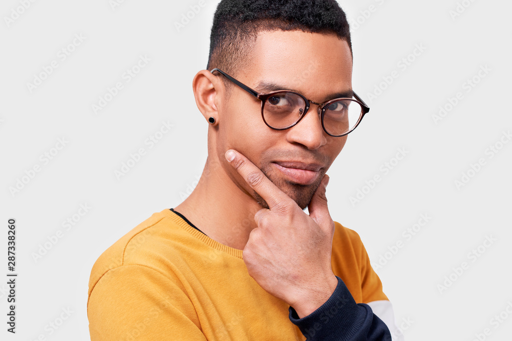 Young African American male wearing sweater and round trandy transparent eyeglasses looking seriously at the camera with pleasant smile, with hand on the chin. People, emotion concept Young African American male wearing sweater and round trandy transparent eyeglasses looking seriously at the camera with pleasant smile, with hand on the chin. People, emotion concept