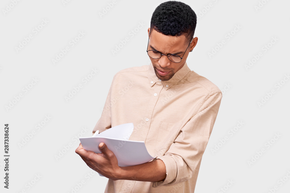 Serious young businessman in casual shirt analyzing financial reports posing on white studio background. African American male student checking paper documents. Serious young businessman in casual shirt analyzing financial reports posing on white studio background. African American male student checking paper documents.