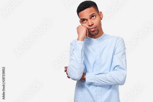 Bored African male leans on hand, has displeased expression, wearing in casual blue shirt, stands against white studio background. Overworked African American man feels tired. People and emotions