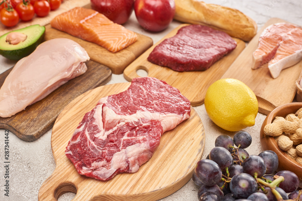 Fresh raw meat, poultry, fish on wooden cutting boards near lemon, grapes, apples, branch of cherry tomatoes, nuts and french baguette on marble surface