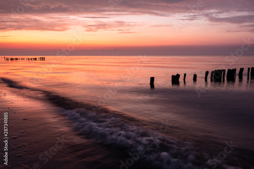 Colorful sunrise at beach of baltic sea in summer with silhouette of old poles groins and wave in foreground, Groemitz, Schleswig-Holstein, Germany photo