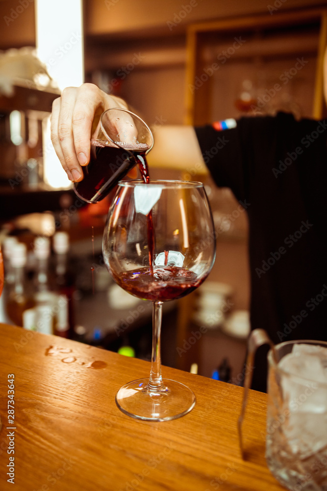 The bartender pours a glass of red wine with bottle in the bar. Glasses of red wine on the bar next to bottles of alcohol