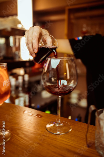 The bartender pours a glass of red wine with bottle in the bar. Glasses of red wine on the bar next to bottles of alcohol