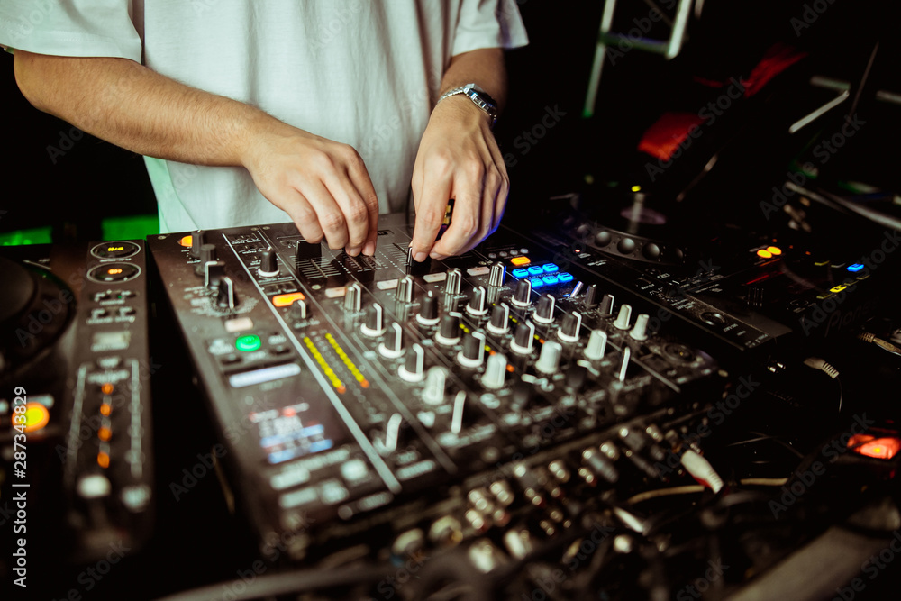Close up view of the hands of a male disc jockey mixing music on his deck with his hands poised over the vinyl record on the turntable and the control switches at night Close up view of the hands of a male disc jockey mixing music on his deck with his hands poised over the vinyl record on the turntable and the control switches at night