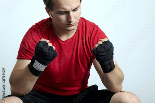 Young caucasian man in sport wear and boxing bandages sits with hands clenched into fists and closed eyes. White wireless earphones, music or pray time. Motivation before fighting. Indoors, copy space