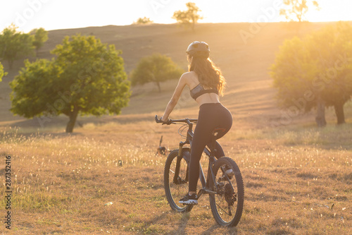 Girl on a mountain bike on offroad, beautiful portrait of a cyclist at sunset, Fitness girl rides a modern carbon fiber mountain bike in sportswear, a helmet, glasses and gloves.