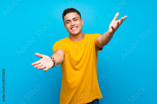 Young man with yellow shirt over isolated blue background presenting and inviting to come with hand