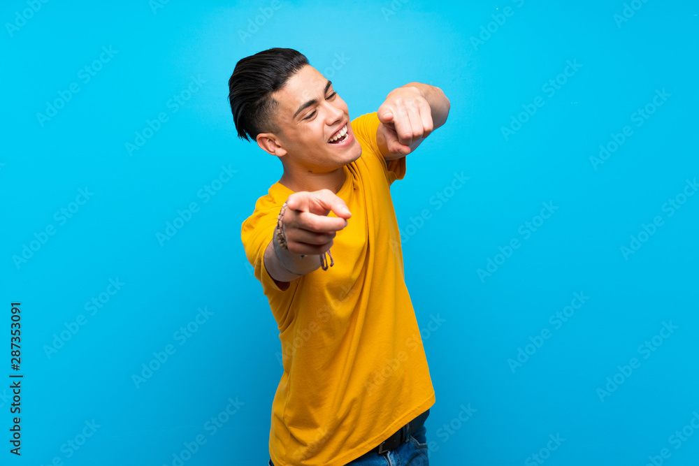 Young man with yellow shirt over isolated blue background points finger at you while smiling Young man with yellow shirt over isolated blue background points finger at you while smiling