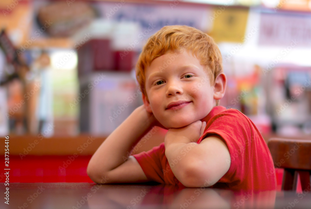 Photo of adorable little boy with ginger hair. Happy child smiling at the camera as he sits at a table resting his chin on his folded arms. Cafe, restaurant Photo of adorable little boy with ginger hair. Happy child smiling at the camera as he sits at a table resting his chin on his folded arms. Cafe, restaurant