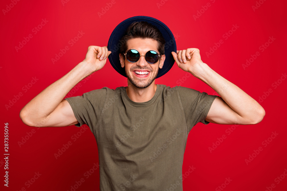 Macho guy holding hands fixing old-fashioned blue hat wear specs grey t-shirt isolated on red background Macho guy holding hands fixing old-fashioned blue hat wear specs grey t-shirt isolated on red background