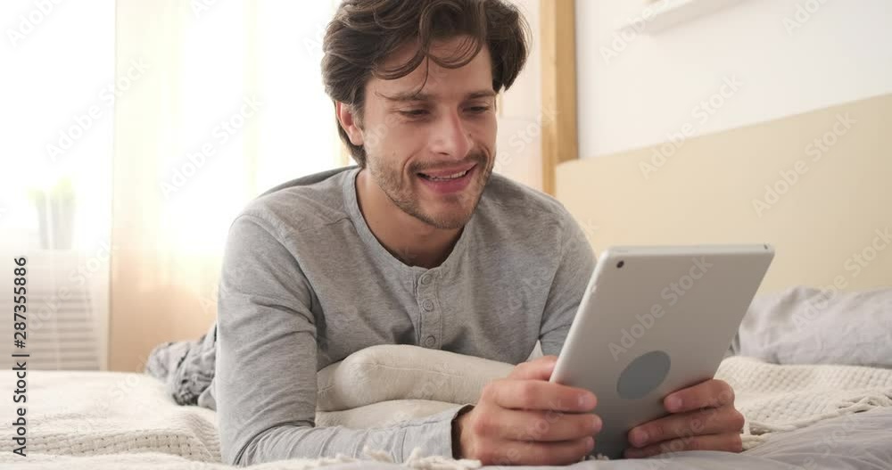 Handsome man in pajamas using digital tablet in his bedroom Handsome man in pajamas using digital tablet in his bedroom