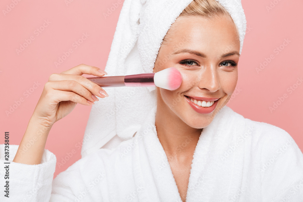 Portrait of caucasian young woman wrapped in white towel after shower applying cosmetics with makeup brush