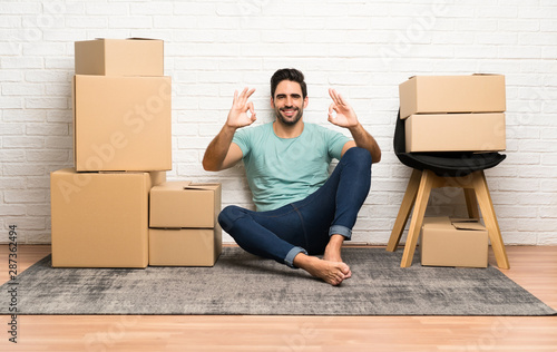 Handsome young man moving in new home among boxes showing an ok sign with fingers