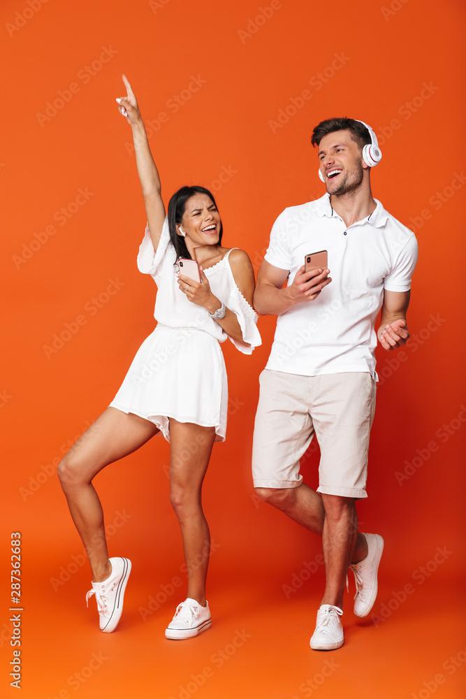 Smiling young amazing loving couple posing isolated over red wall background using mobile phones listening music with headphones.