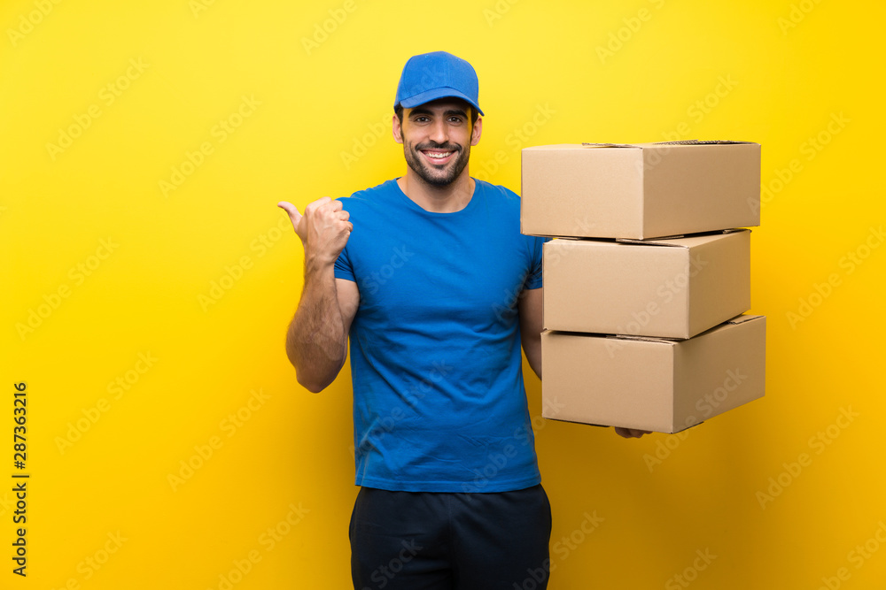 Young delivery man over isolated yellow wall pointing to the side to present a product Young delivery man over isolated yellow wall pointing to the side to present a product