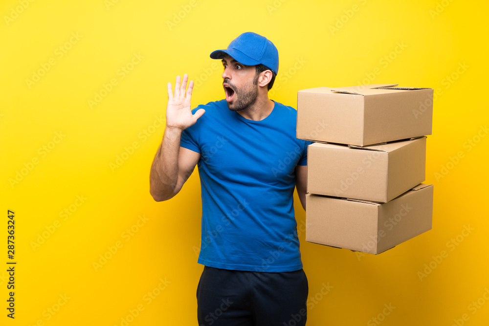 Young delivery man over isolated yellow wall shouting with mouth wide open Young delivery man over isolated yellow wall shouting with mouth wide open