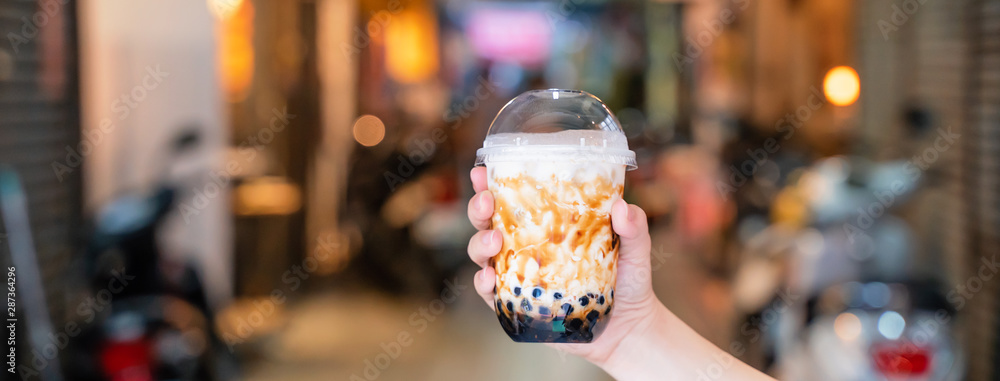 © RomixImage - Young girl is holding and showing a cup of brown sugar flavored tapioca pearl bubble milk tea in night market of Taiwan background, close up, bokeh