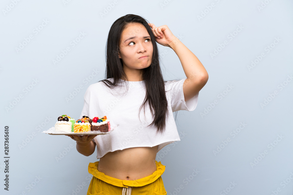 Teenager asian girl holding lots of different mini cakes having doubts and with confuse face expression Teenager asian girl holding lots of different mini cakes having doubts and with confuse face expression