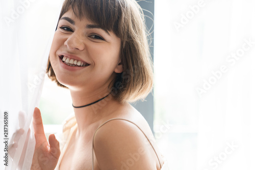 Happy cheery beautiful young woman posing indoors at home near window with curtain. Free Download Happy cheery beautiful young woman posing indoors at home near window with curtain. - GFXLoad