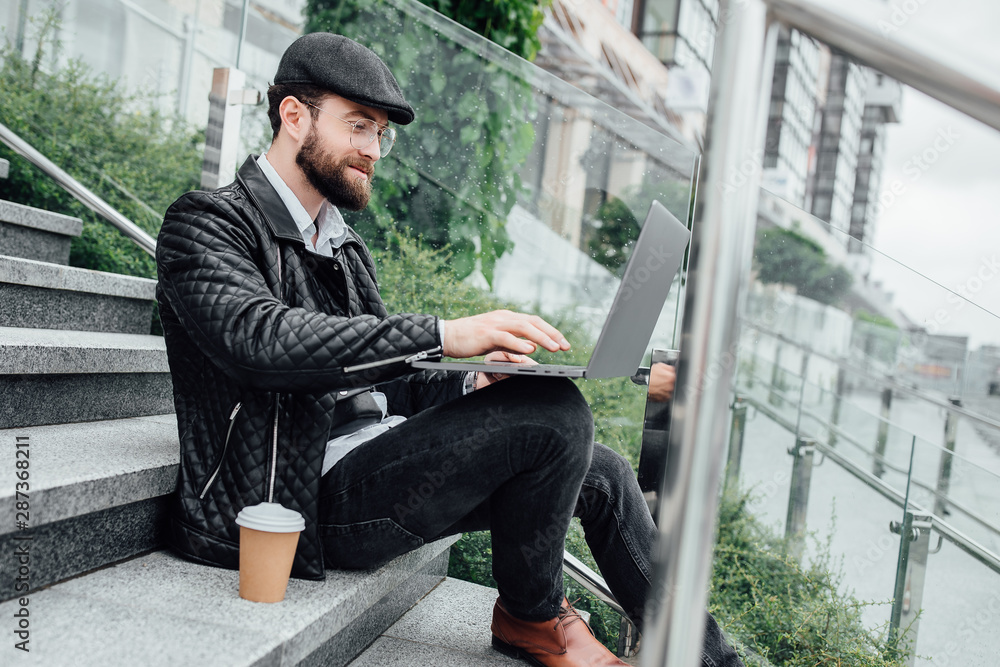 Handsome smiling manager man drinking coffee and working on laptop while sitting outdoors on the stairs. Business concept. Handsome smiling manager man drinking coffee and working on laptop while sitting outdoors on the stairs. Business concept.