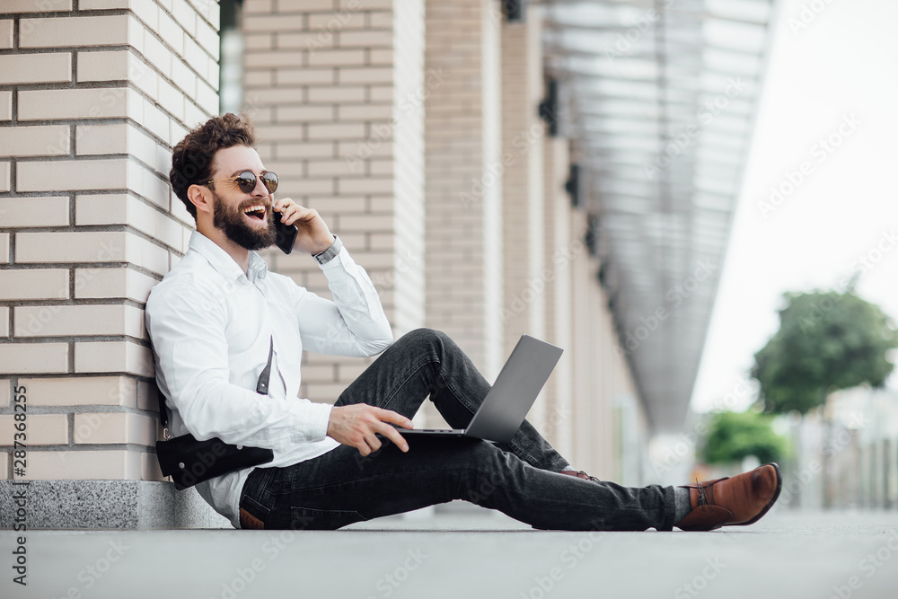A bearded, happy, smiling, stylish man sitting on the flour on the streets of the city near modern office centre and workin with his laptop and calling phone. Frank laugh. A bearded, happy, smiling, stylish man sitting on the flour on the streets of the city near modern office centre and workin with his laptop and calling phone. Frank laugh.
