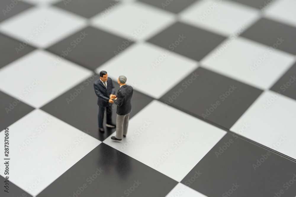 Miniature 2 people businessmen Shake hands standing on a chessboard with a chess piece on the back Negotiating in business. as background business concept and strategy concept with copy space. Miniature 2 people businessmen Shake hands standing on a chessboard with a chess piece on the back Negotiating in business. as background business concept and strategy concept with copy space.