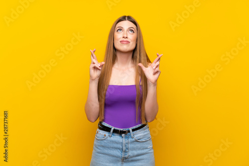 Young woman over isolated yellow background with fingers crossing and wishing the best