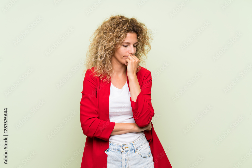Young blonde woman with curly hair over isolated green background having doubts Young blonde woman with curly hair over isolated green background having doubts