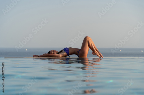 Young female in bikini lying by infinity swimming pool on a summer day. Woman tanning poolside with sea in background.