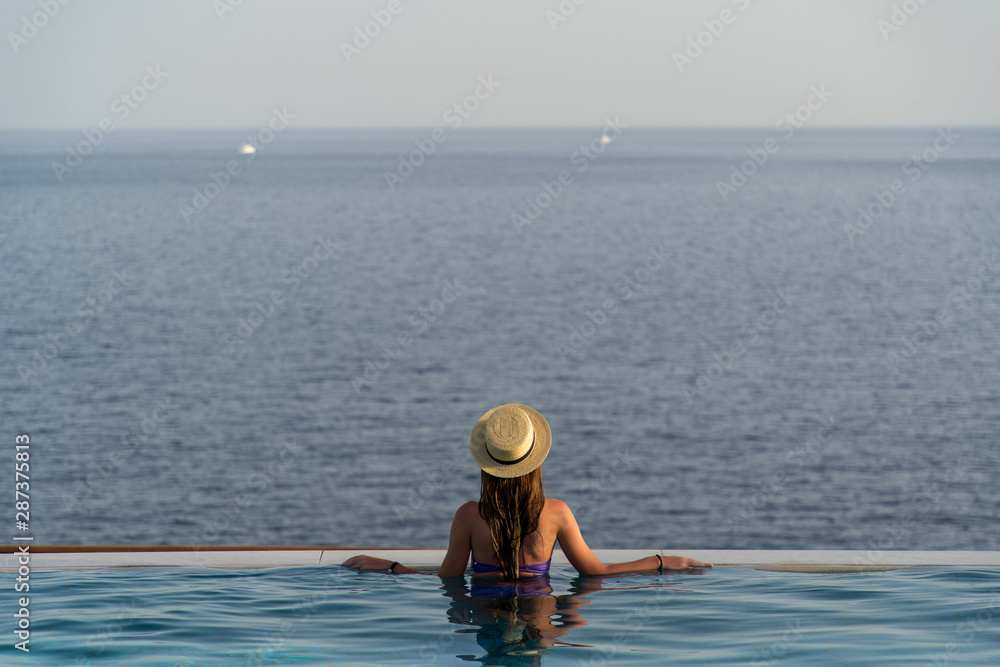 Beautiful young woman with a hat is relaxing in an infinity pool with a breathtaking view