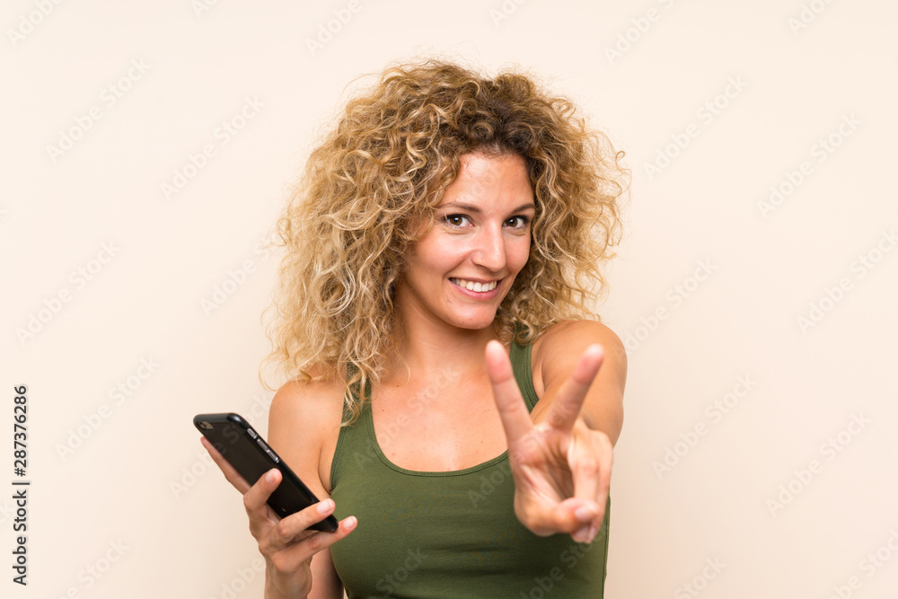 Young blonde woman with curly hair using mobile phone smiling and showing victory sign Young blonde woman with curly hair using mobile phone smiling and showing victory sign