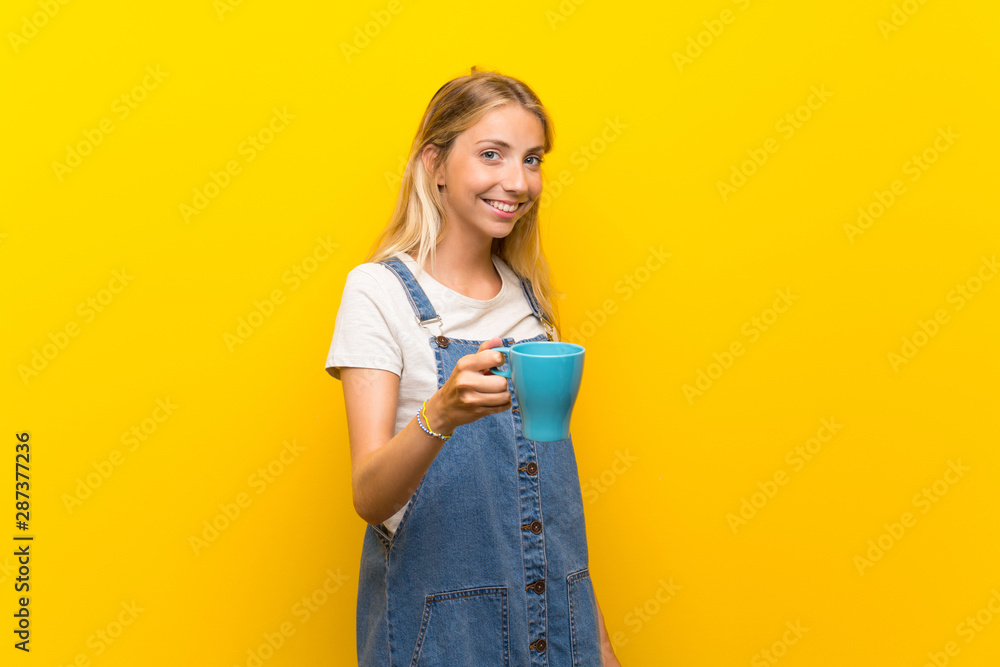 Blonde young woman over isolated yellow background holding hot cup of coffee Blonde young woman over isolated yellow background holding hot cup of coffee