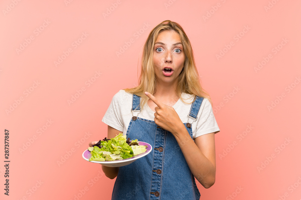 Blonde young woman with salad over isolated pink wall surprised and pointing side